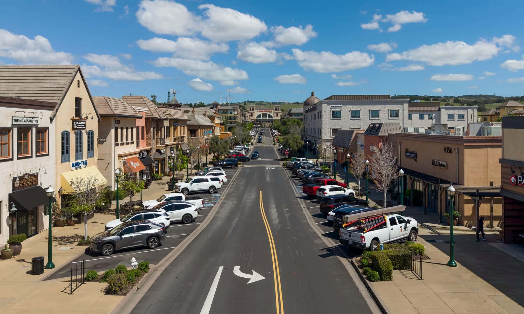 The main street of El Dorado Hills Town Center — a wide two-lane road, angle parking on both sides, tile-roofed shopfronts and the dry foothills behind