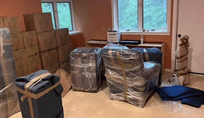 A living room packed up: armchairs shrink-wrapped over moving blankets, a floor lamp padded, blanket bundles strapped, cartons stacked along the wall