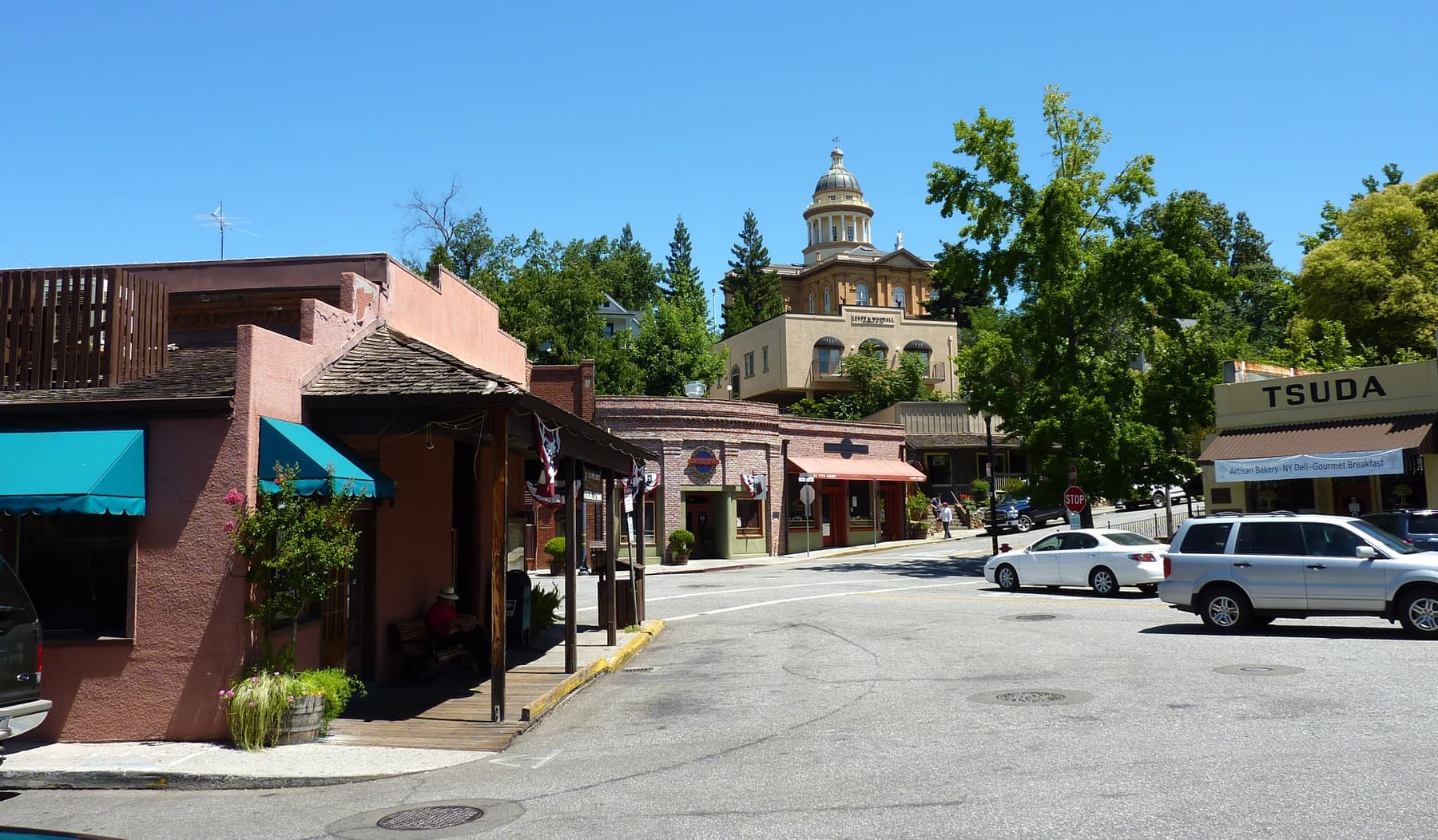 A street corner in Old Town Auburn — a stucco storefront with awnings and a timber porch, cars at the kerb, and the domed Placer County Courthouse on the wooded hill above the town