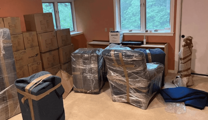 A living room packed up: armchairs shrink-wrapped over moving blankets, a floor lamp padded, blanket bundles strapped, cartons stacked along the wall