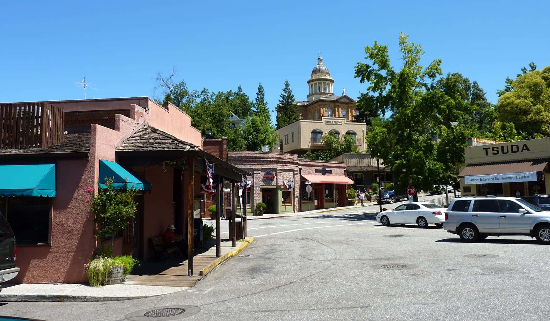 A street corner in Old Town Auburn — a stucco storefront with awnings and a timber porch, cars at the kerb, and the domed Placer County Courthouse on the wooded hill above the town